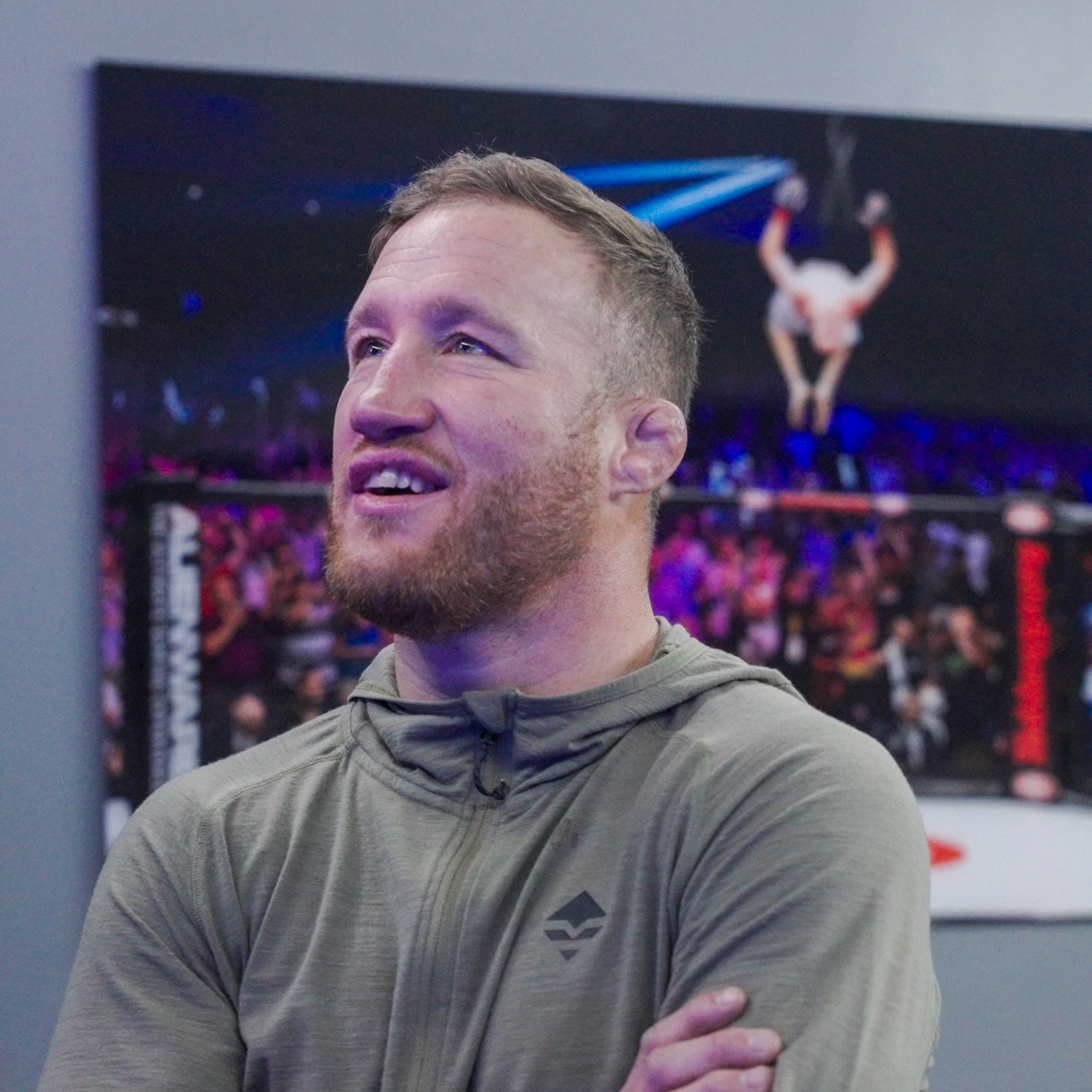 Gaethje smiling in an olive hoodie, fight imagery on the LED wall behind him